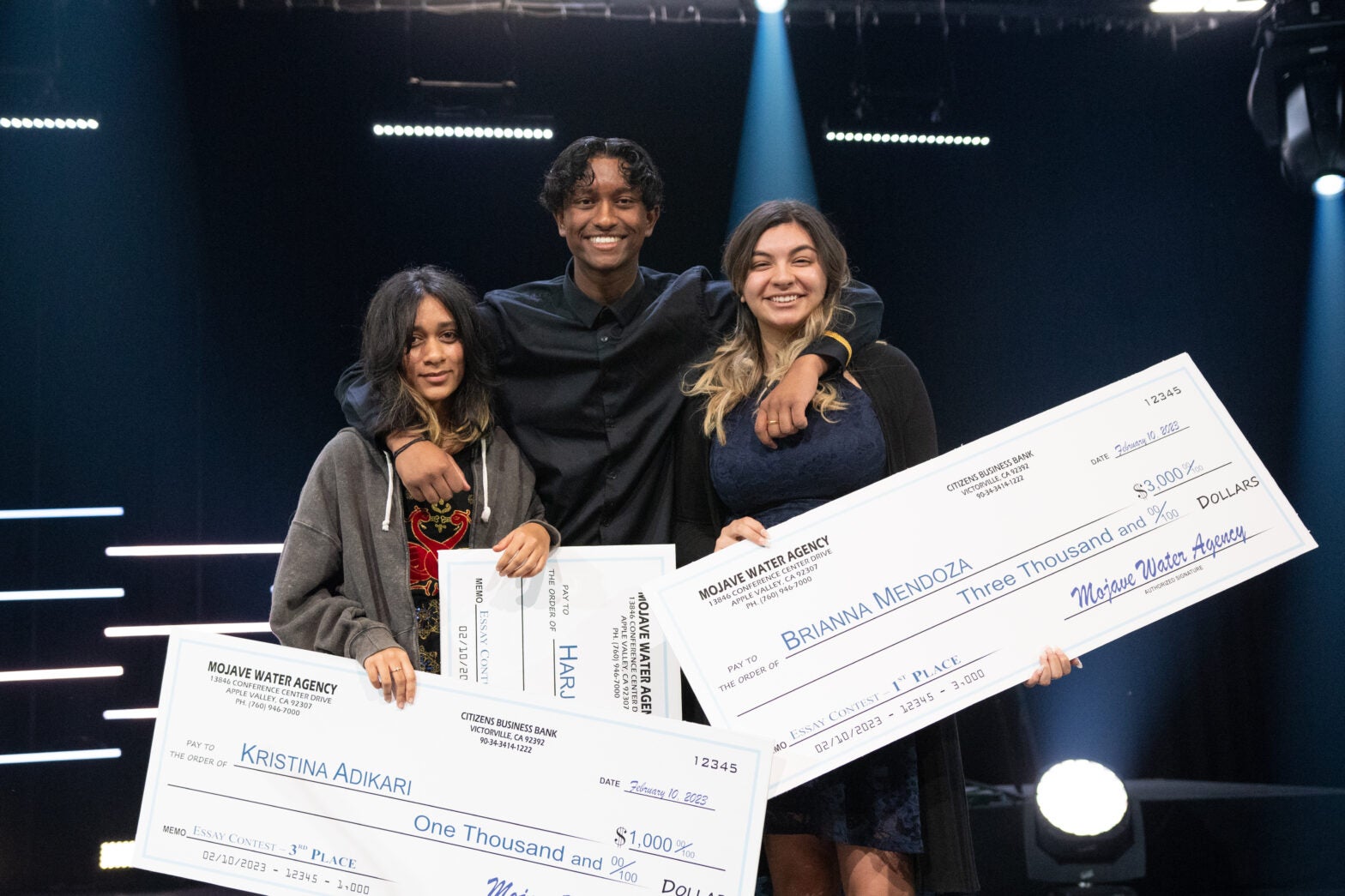 Three young people stand on stage holding oversized checks, smiling at the camera with stage lights in the background.