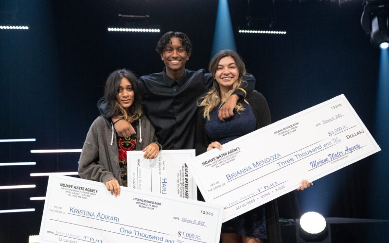 Three young people stand on stage holding oversized checks, smiling at the camera with stage lights in the background.
