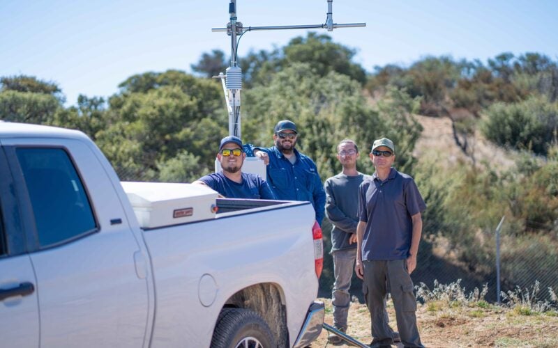 Four men stand next to a white pickup truck and a weather station sensor in an outdoor, brushy area under a clear sky.