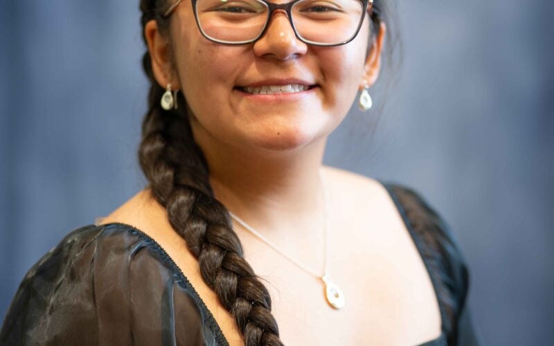 Young woman with long dark hair in a braid, wearing glasses, a black sheer-sleeve dress, and jewelry, smiles in front of a blurred blue-gray background.