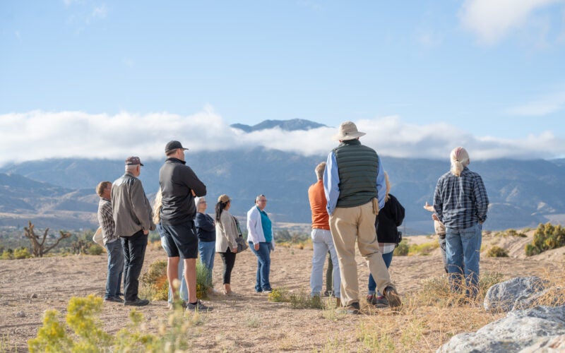 A group of people stands outdoors on rocky terrain, facing a mountain range in the distance under a partly cloudy sky.