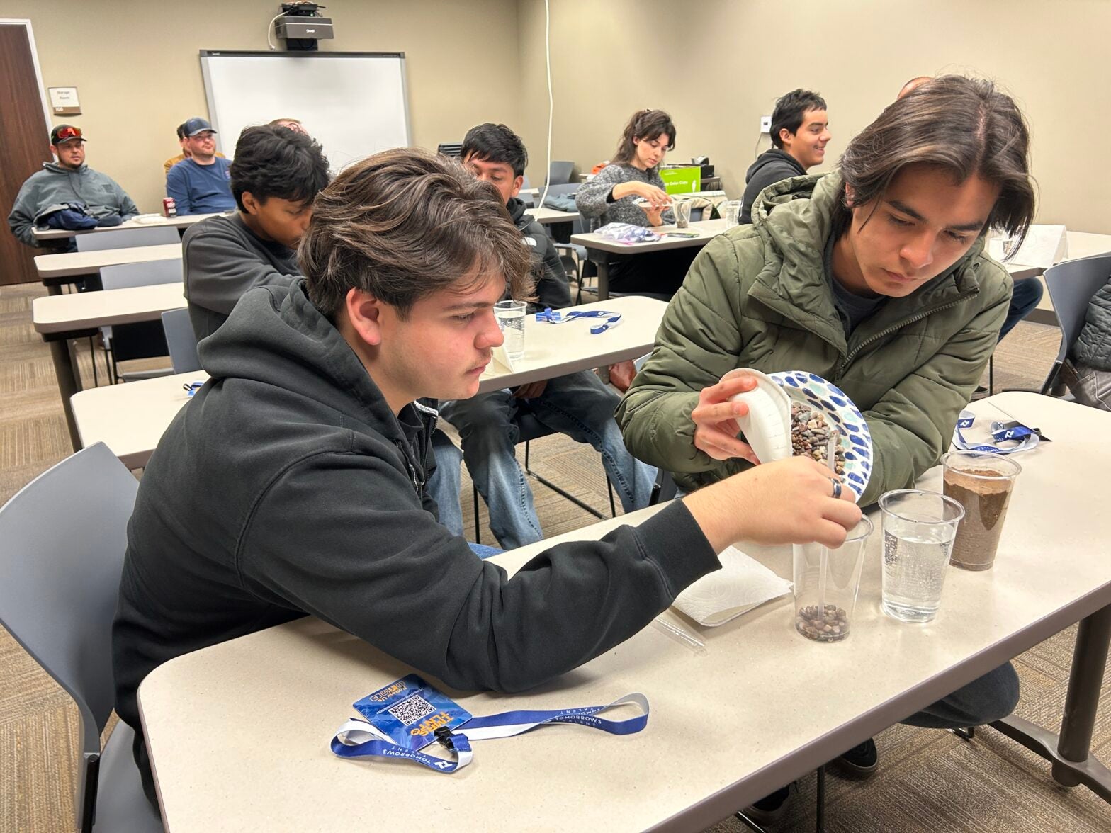 Two young men sit at a classroom table, pouring small stones from a plate into a clear cup during a group activity. Other participants and materials are visible in the background.