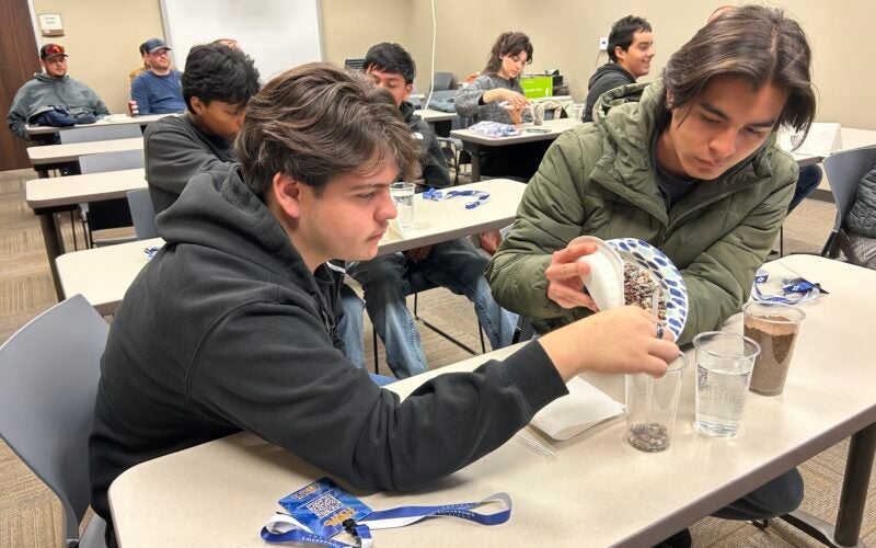 Two young men sit at a classroom table, pouring small stones from a plate into a clear cup during a group activity. Other participants and materials are visible in the background.