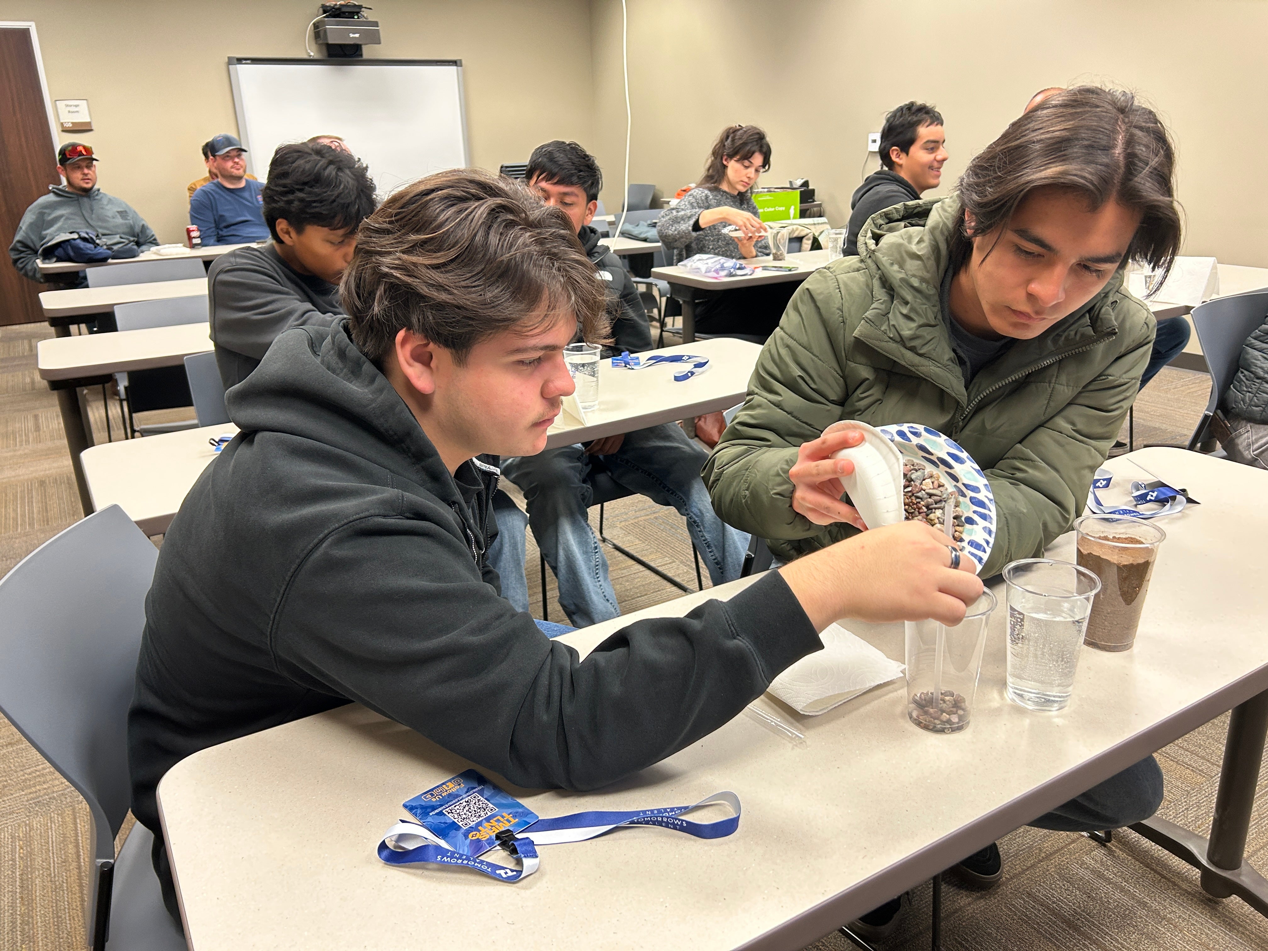 Two young men sit at a classroom table, pouring small stones from a plate into a clear cup during a group activity. Other participants and materials are visible in the background.