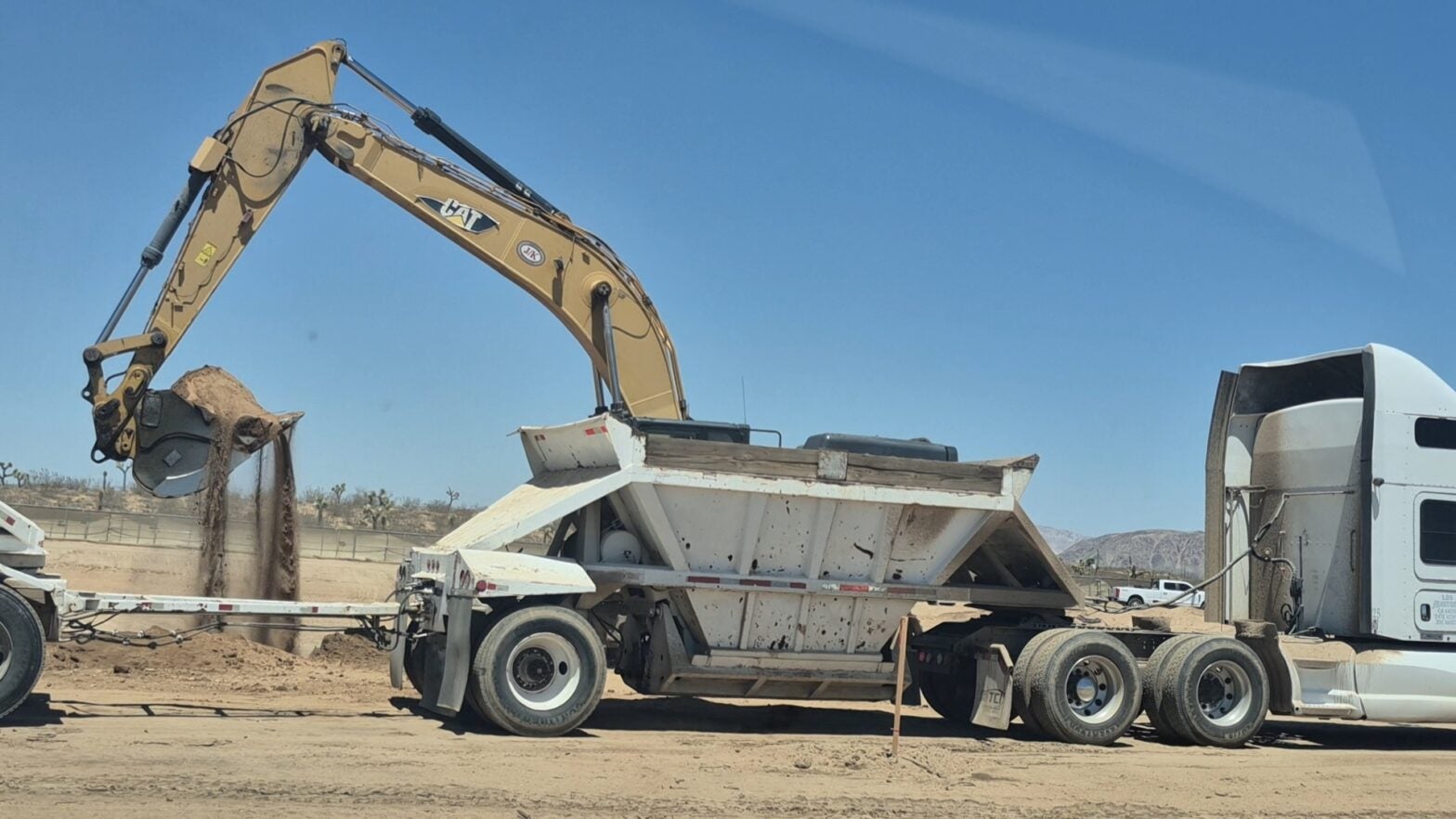 An excavator loads dirt into the back of a white dump trailer attached to a semi-truck on a construction site under a clear sky.