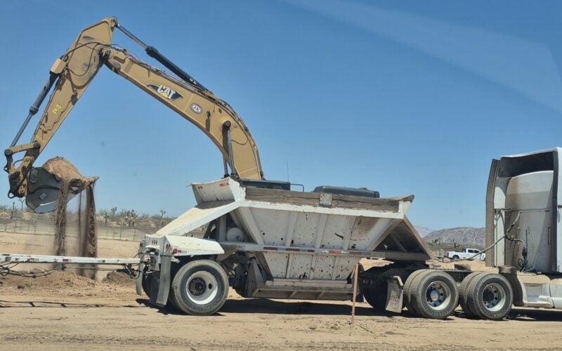 An excavator loads dirt into the back of a white dump trailer attached to a semi-truck on a construction site under a clear sky.