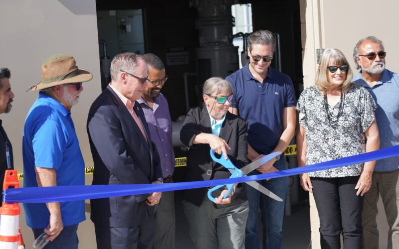 A group of people stands by as an older woman uses large scissors to cut a blue ribbon at a building entrance during a ribbon-cutting ceremony.
