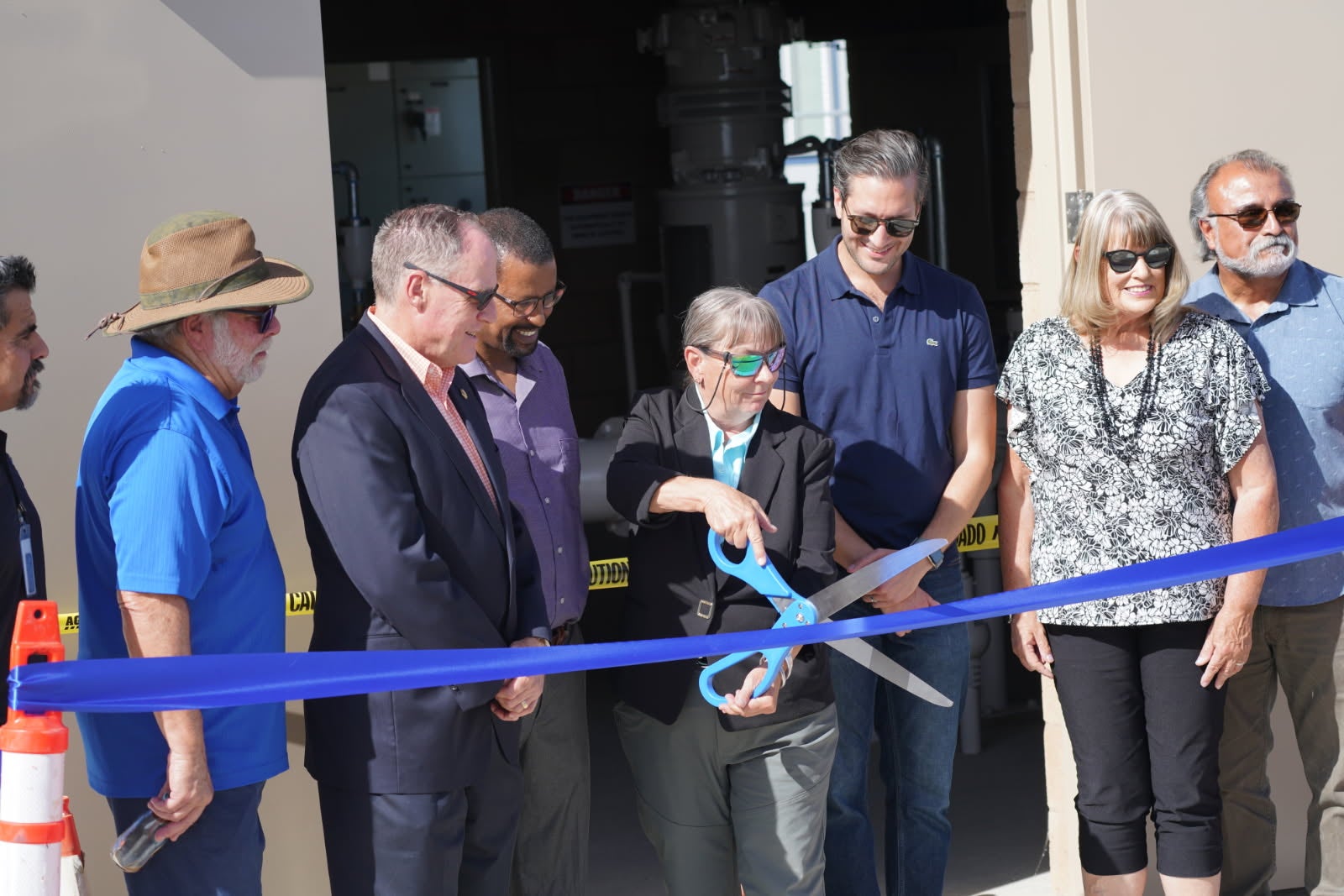 A group of people stands by as an older woman uses large scissors to cut a blue ribbon at a building entrance during a ribbon-cutting ceremony.