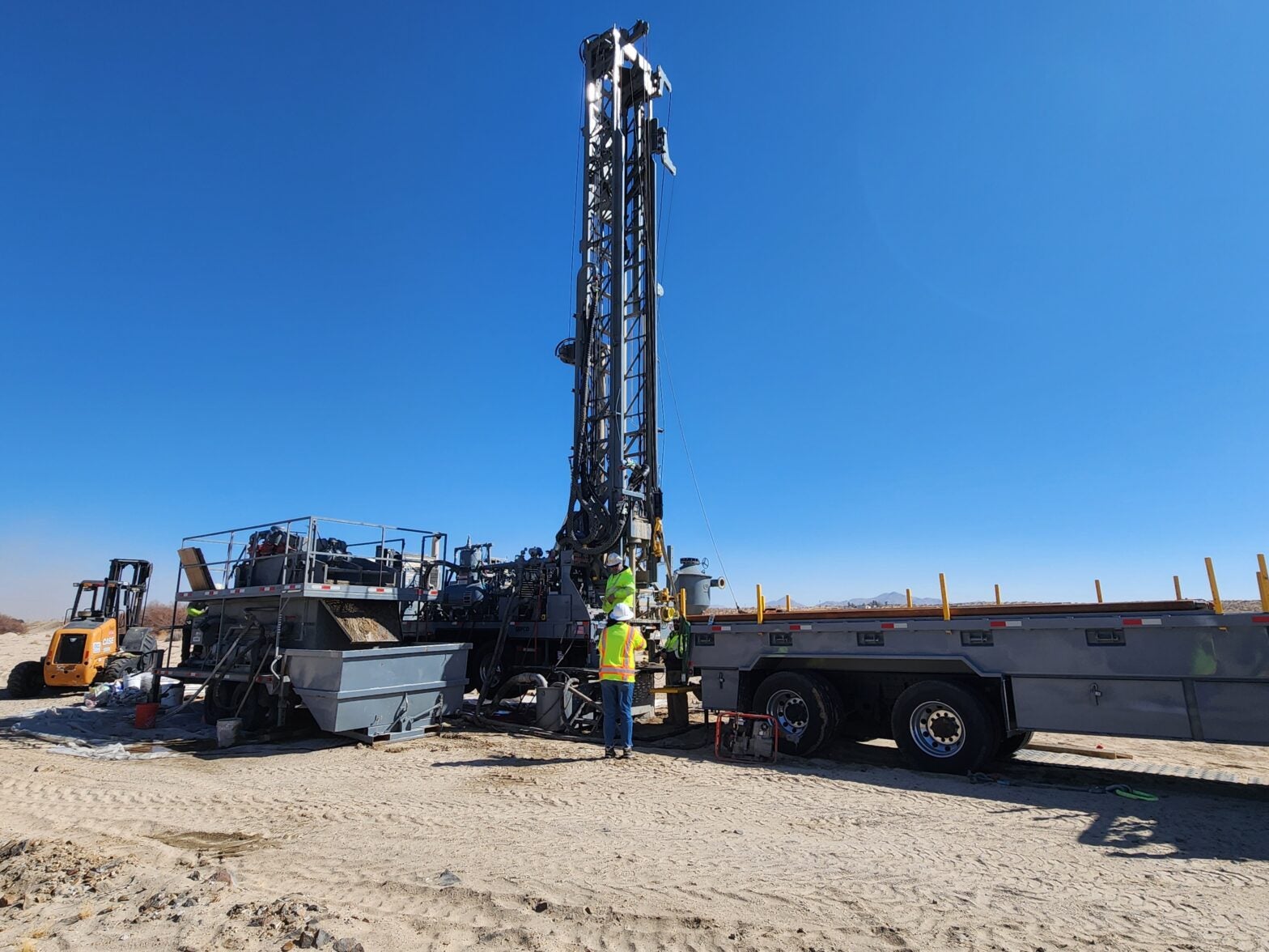 Two workers in high-visibility vests operate a large drilling rig mounted on a flatbed trailer in a sandy area.