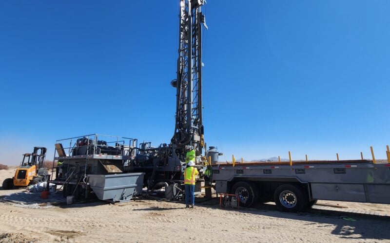 Two workers in high-visibility vests operate a large drilling rig mounted on a flatbed trailer in a sandy area.