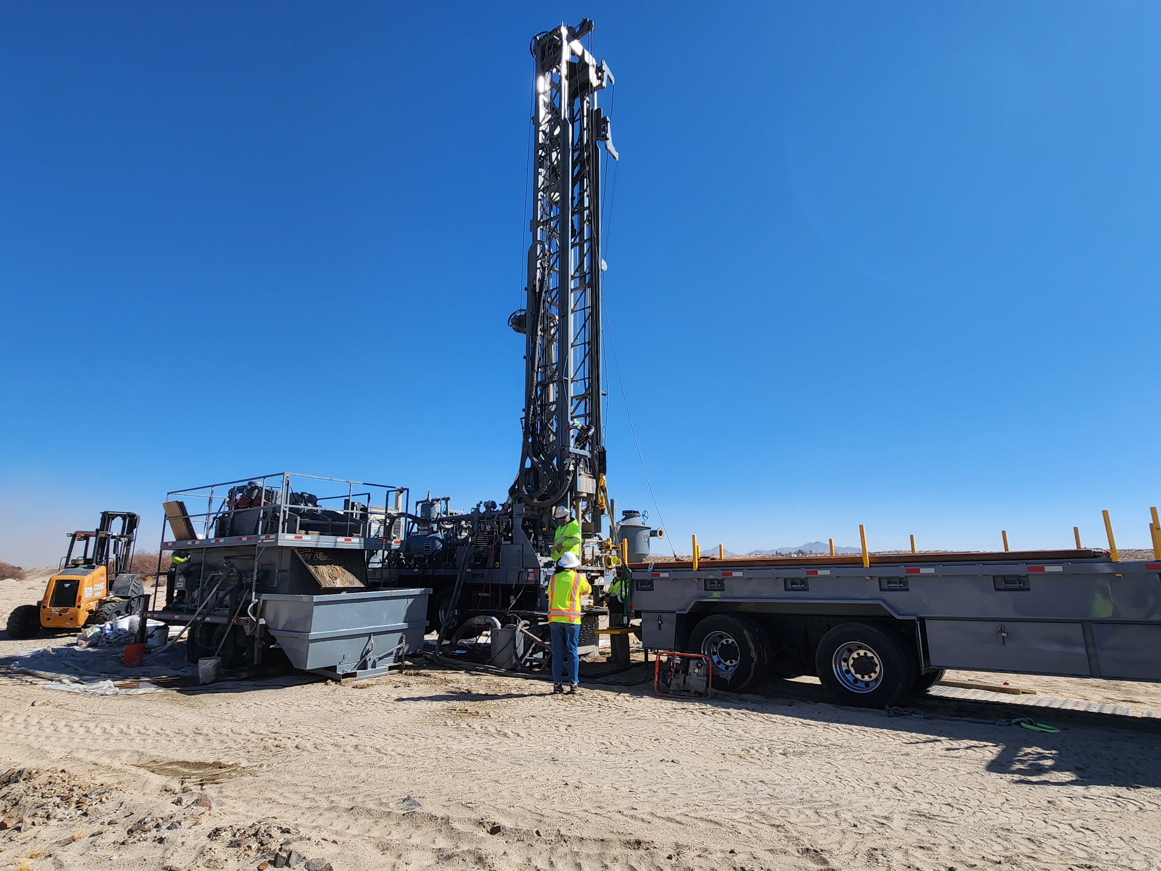 Two workers in high-visibility vests operate a large drilling rig mounted on a flatbed trailer in a sandy area.